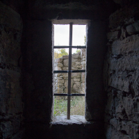 An ancient stone window frame, illuminated by natural light, opens up to a view of crumbling walls and overgrown vegetation in Saaremaa, encapsulating a sense of historical decay and the resilience of