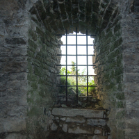 Ancient stone-walled alcove with a barred window on the island of Saaremaa, Estonia, overlooking lush foliage, evoking a sense of historical confinement and the contrast between human structures and