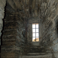 A dimly lit stone spiral staircase winding up to a narrow window with iron bars, within the weathered walls of an ancient structure on Saaremaa Island, Estonia.