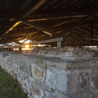 Under a rustic bridge with a lattice of metal beams in Saaremaa, the sun sets in the background, casting a warm glow and creating a play of light and shadow on the stone base and