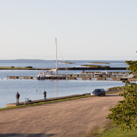 A serene lakeside scene on the Estonian island of Saaremaa features a small harbor with a sailboat docked at the pier. Two people stand along the shoreline near parked bicycles, while another person is standing by a parked car on a gravel path. Calm waters of the Baltic Sea and distant small islands under a clear sky complete the peaceful setting.