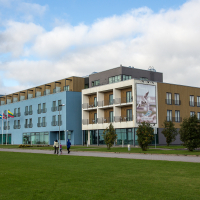 A modern educational building with colorful façades, displaying flags and a banner, under a cloudy sky, with pedestrians walking by on a lush green lawn in Saaremaa.