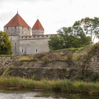A medieval stone fortress with conical red-tiled roofs stands majestically beside a serene river on Saaremaa, surrounded by lush greenery and remnants of ancient walls under a cloudy sky.
