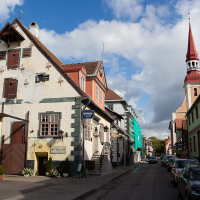 A quaint Saaremaa street lined with colorful, traditional buildings under a blue sky with fluffy white clouds, showcasing architecture from different periods and a tall church spire in the distance.