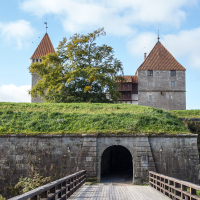 Medieval stone fortress in Saaremaa with twin towers atop a grassy mound, under a clear blue sky, accessible via a wooden bridge leading to an arched entrance.