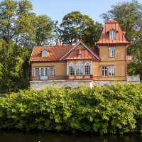A picturesque, two-story house with ornate wooden architecture and topped with red roofs stands proudly in Saaremaa. Surrounded by lush greenery, the building features detailed windows and a stone foundation. A serene body of water sits in the foreground, while tall, leafy trees frame the background.