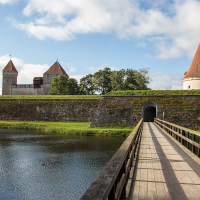 A serene wooden bridge extends over tranquil waters toward an ancient fortress with conical towers in Saaremaa, nestled under a blue sky with clouds.