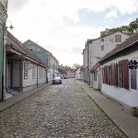 A quiet cobblestone street on Saaremaa Island, lined with quaint, low-rise buildings under a cloudy sky, with a car parked in the distance and signs suggesting commercial activity.