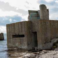 Ruins of a coastal military bunker in Saaremaa, eroded by time and the sea, under a cloud-dappled sky, with remnants of its walls standing testament to a bygone