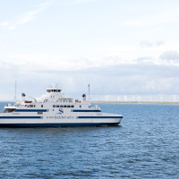 A white passenger ferry traverses calm blue waters towards Saaremaa island, under a mostly clear sky, with a wind farm visible on the horizon, showcasing sustainable energy alongside maritime travel.