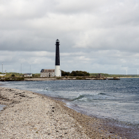 A solitary black and white lighthouse stands guard on Saaremaa's rugged coastline under a cloudy sky, with gentle waves lapping at the stony shore, hinting at a blend of tranqu