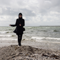 A person in a black coat and beret stands on the pebbly shore of Saaremaa Island, gazing towards the camera, with turbulent sea waves and a cloudy sky in the background
