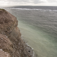 A rugged cliff on Saaremaa Island overlooks a turbulent sea under a cloud-covered sky, with waves lapping against the rocky shore, highlighting the dynamic interface between land and water.