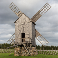An old wooden windmill with weathered sails stands atop a stone base in Saaremaa, set against a cloudy sky, with a fenced green field in the backdrop.