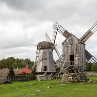 An old wooden windmill with weathered sails stands in a pastoral Saaremaa landscape, surrounded by green grass and a rustic stone wall under a cloudy sky.