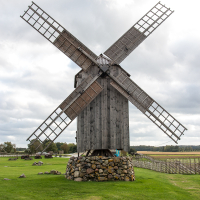 A traditional wooden windmill with a stone foundation stands under an overcast sky on a lush green lawn in Saaremaa, surrounded by a wooden fence and scattered boulders.