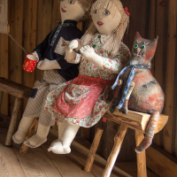 Handcrafted fabric dolls depicting a couple and a cat seated on a wooden bench, with the male doll holding a red cup and the female doll adorned with a flower in her hair, capturing a homely