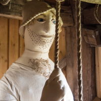 A lifelike mannequin wearing a medieval-style hood and tunic stands indoors in Saaremaa, its hand gripping a thick rope, with a contemplative expression on its face, surrounded