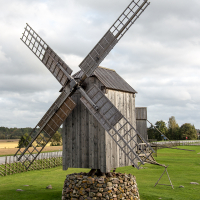A traditional wooden windmill stands on a stone base amidst a grassy field in Saaremaa, its four large sails at rest beneath a cloudy sky, evoking a pastoral serenity in a