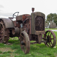 An old, rusted tractor stands idle on the grass in Saaremaa, showcasing its vintage design and iron wheels, with a traditional wooden fence and cloudy sky in the background, evoking nostalgia