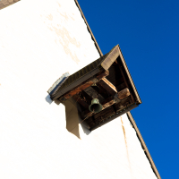 A bell visible through an open wooden attic window on a stark white wall under a deep blue sky, hinting at a traditional Saaremaa structure with a minimalist architectural style.
