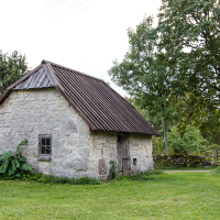 An old stone cottage with a tiled roof, surrounded by lush greenery and a wooden fence, stands quietly in the serene Saaremaa countryside landscape.