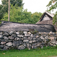 A rustic stone wall foregrounds traditional wooden buildings with thatched roofs, nestled in the tranquil green landscape of Saaremaa, exuding old-world charm and historical Scandinavian architecture.