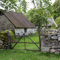 An old stone cottage with a corrugated metal roof, surrounded by a rustic stone wall and a wooden gate, nestled amid lush greenery in Saaremaa, evokes the charm of rural