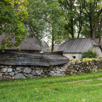 An overturned wooden boat rests atop a stone wall, with quaint thatched-roof cottages nestled among green trees in the serene, rustic village setting of Saaremaa.