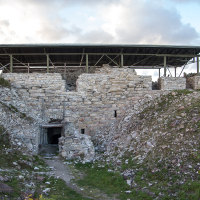 An ancient stone structure on Saaremaa Island with weathered walls sits under a modern protective canopy, preserving its historical importance, with a pathway leading to a darkened entrance, evoking intrigue and