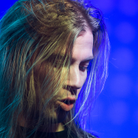 A person with long, flowing hair and a contemplative expression is illuminated by vibrant blue and green stage lights, creating a dramatic effect. Reminiscent of a Sabaton concert, they are looking downward with their hair partially covering their face, set against a blurred background.