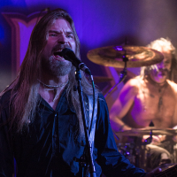 A male musician with long hair and a beard is singing passionately into a microphone while playing a guitar on stage. Wearing a dark shirt, he channels the energy of Sabaton. In the background, a shirtless drummer is intensely playing the drums under stage lights, creating a dynamic band atmosphere.
