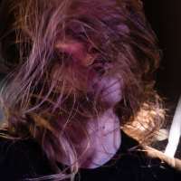 A person with long, blonde hair is captured in motion, with hair wildly obscuring their face. The individual, clad in a black shirt, seems to be caught in an intense moment, possibly performing live with the band Sabaton. The dark background adds to the dynamic and energetic atmosphere.