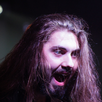 A bearded person with long brown hair is enthusiastically performing on stage, with a guitar strap visible over their right shoulder. The dim purple lighting highlights their animated expression, with mouth open and eyes wide, conveying a sense of excitement or passion characteristic of a heavy metal band like Sabaton.
