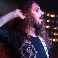 A bearded man with long hair and a mustache plays a guitar. He is wearing a black shirt with a Sabaton lion crest design. His hand is raised to his ear, and stage lights with a warm glow are visible in the background.