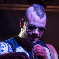 A person with a mohawk hairstyle, wearing sunglasses and a sleeveless top with metallic shoulder plates, holds a microphone and appears to be performing on stage. A visible tattoo can be seen on the person's right arm, and stage lighting illuminates the scene, reminiscent of a Sabaton concert.
