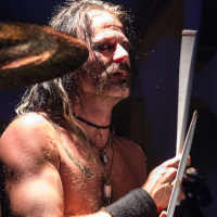 A shirtless drummer from a Swedish heavy metal band, with long, wet hair, is captured mid-performance. He is holding drumsticks and adorned with necklaces, including one with a large pendant. He also wears a black armband on his right forearm. The lighting focuses on him, highlighting the intensity of his expression.