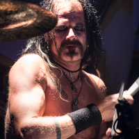 A drummer with long black hair and a mustache is playing the drums under stage lighting. He is shirtless, revealing muscular arms with black wristbands and a tattoo on his left arm. His expression is focused, and he holds a drumstick in his right hand, immersed in the powerful music of Sabaton.