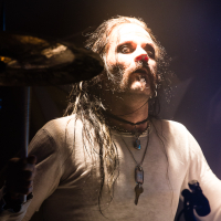A drummer with long wet hair and a moustache passionately plays, illuminated by dramatic lighting. Part of the band Sabaton, he wears a light-colored long-sleeve shirt and multiple necklaces, including a cross pendant. One drumstick is raised while the other is mid-motion, captured in this intense music moment against a dark background.