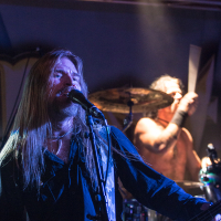 A long-haired musician sings passionately into a microphone on stage. He is dressed in dark clothing and lit dramatically with stage lights. Behind him, a shirtless drummer plays energetically on a drum kit, also illuminated by stage lighting. Both are performing in a concert setting for the renowned metal band Sabaton.