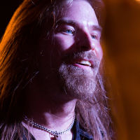 A bearded man with long hair smiles while looking slightly to the side. He is wearing a necklace with a fang-like pendant and a leather vest, evoking a rocker vibe. The background is dimly lit, highlighting his face and expression's details, as if he's about to take the stage with Sabaton.