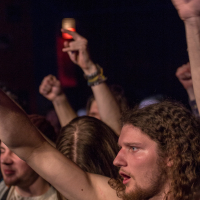 An enthusiastic crowd at a heavy metal concert with arms raised. One person with long curly hair is at the forefront, making the "rock on" hand gesture, while fans around them cheer for Sabaton, capturing the energetic and lively atmosphere of the band's performance.