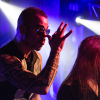 Two male singers on stage during a live concert. The front singer from the Swedish power metal band Sabaton, wearing sunglasses and a sleeveless shirt, reveals his tattooed arms while making a gesture with his hand. The other singer beside him has long hair and a beard. The background features vibrant lighting with a blue and purple hue.