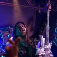 A long-haired musician passionately holds up a white and black electric guitar on stage, illuminated by colored spotlights, evoking the spirit of Sabaton. Wearing a black outfit and mid-performance, he electrifies the audience, whose raised hands capture the energetic atmosphere of the concert.