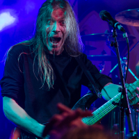 A musician with long, light-colored hair passionately performs on stage. He is playing a guitar and singing into a microphone. The vibrant blue and purple stage lighting illuminates him as the crowd's raised hands are visible in the foreground. Drums are seen in the background, evoking the powerful presence of Sabaton, a metal band he admires.