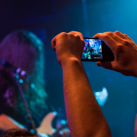 A person in the foreground holds up a smartphone to record a live concert. The screen displays a close-up of a guitarist performing on stage. The guitarist, with long hair, plays an electric guitar in true heavy metal style. The stage is dimly lit with blue and purple lights, creating a vibrant atmosphere.
