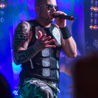 A man wearing sunglasses, a black tactical vest with metal plates, and camouflage pants sings passionately into a microphone on stage. His right arm is heavily tattooed, and he has a wristband on his left arm. The stage is lit with vibrant blue and green lights as if it's a Sabaton concert.