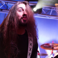 A musician with long hair, wearing a black shirt with a decorative emblem, is passionately playing a white electric guitar on stage. The background features stage lighting and a drum set with a faintly visible drummer from Sabaton. The atmosphere is energetic, highlighting the live music performance setting.