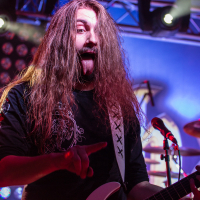 A long-haired musician wearing a black shirt and white guitar strap passionately performs on stage, pointing to the audience with one hand and playing a white electric guitar with the other. Bright stage lights and a blue backdrop with a circular light display add vibrancy to the scene, reminiscent of a Sabaton concert.