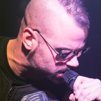 A person with a mohawk hairstyle and wearing sunglasses is performing on stage, leaning into a microphone. They are wearing a sleeveless outfit with a metal plate design on the shoulder, reminiscent of the band Sabaton. The background is blurred and hazy, likely from stage lighting, indicating a concert setting.